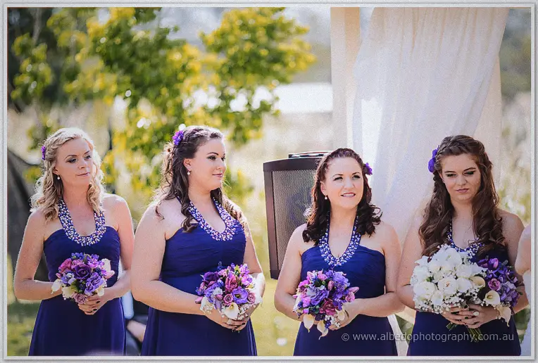Four bridesmaids in violet halter gowns hold purple bouquets at a sunlit garden ceremony at Brookleigh Estate