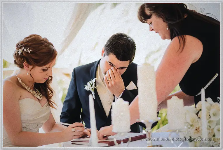 Groom wipes away tears as Amanda signs the register beside him in a candlelit marquee at Brookleigh