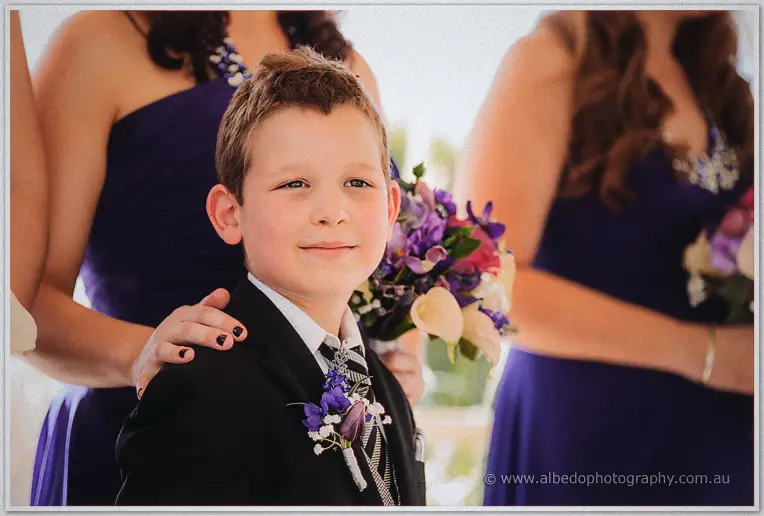 A young page boy in a black suit with a purple boutonniere smiles between purple-gowned bridesmaids