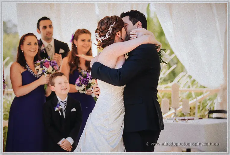 Craig kisses his bride as smiling bridesmaids and the young page boy look on under white draping