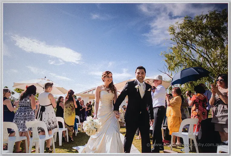 Amanda and Craig walk hand in hand back down the aisle as guests cheer beneath a blue summer sky