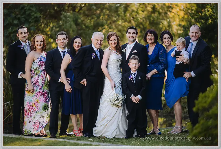 The full wedding party poses on the lawn, bride holding white bouquet flanked by family