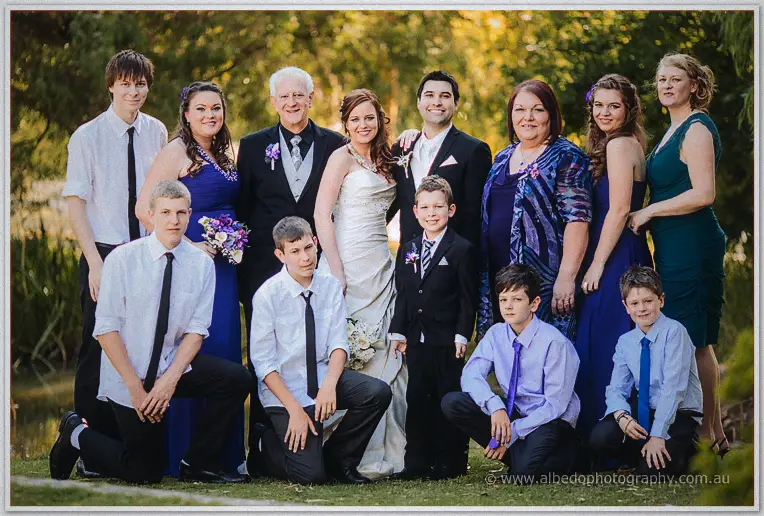 Extended family group with kids in shirts and ties around the newlyweds beside a golden pond