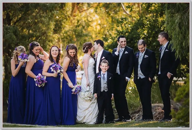 Craig kisses Amanda's cheek as giggling bridesmaids and groomsmen line the lakeside path