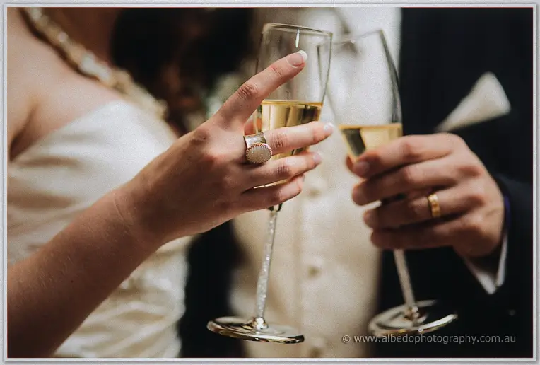 Close-up of bride and groom clinking champagne flutes, rings catching the candlelight