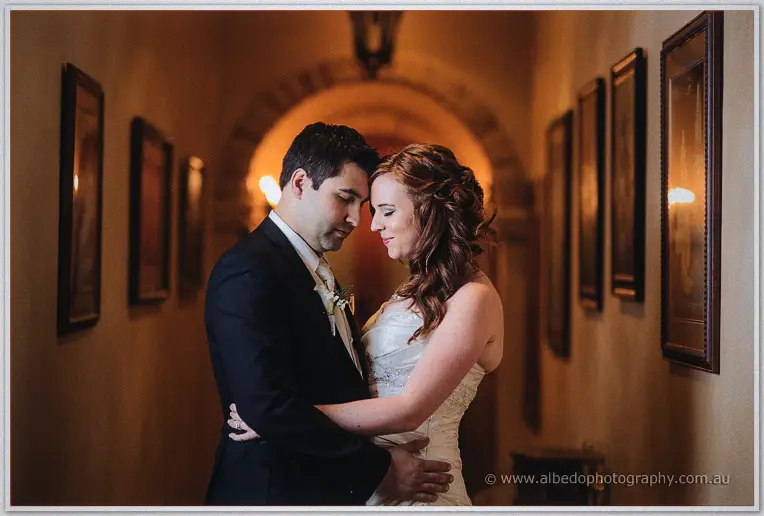 The couple embrace in a lantern-lit homestead hallway lined with framed paintings