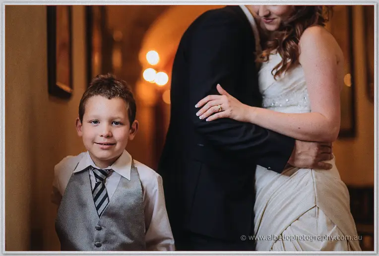 A grinning page boy in grey vest stands before Amanda and Craig embracing in the corridor