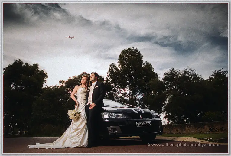Amanda and Craig lean on a black sedan beneath a moody sky with a jet overhead
