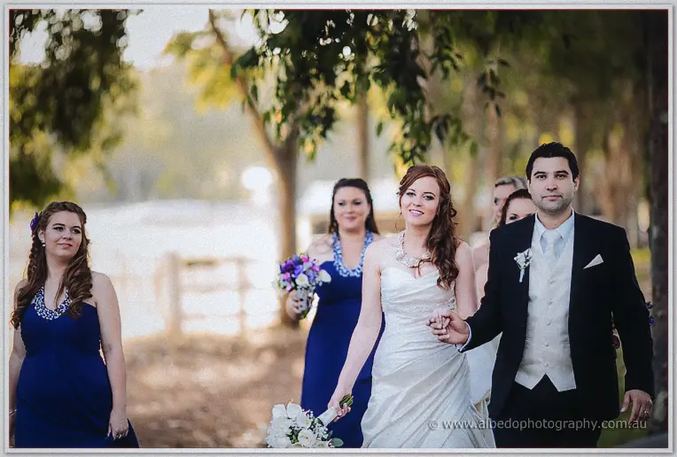Amanda and Craig walk arm in arm past bridesmaids along the sunlit paddock fence