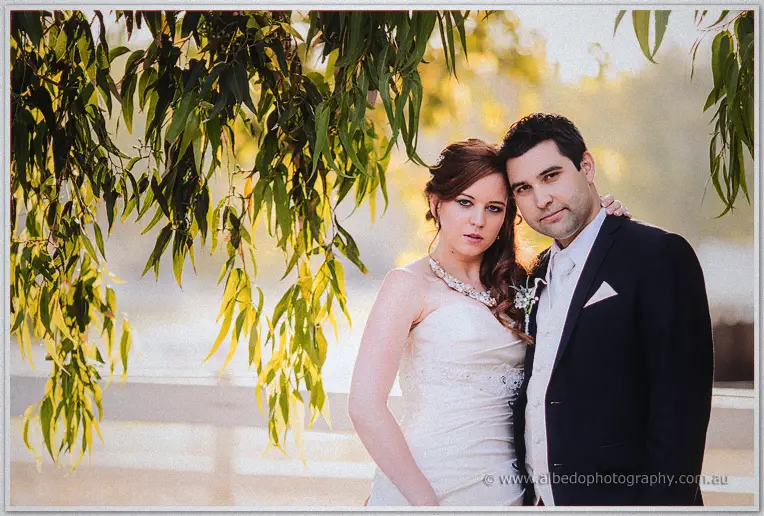 Craig holds Amanda close under willow leaves by the water, both gazing at the camera