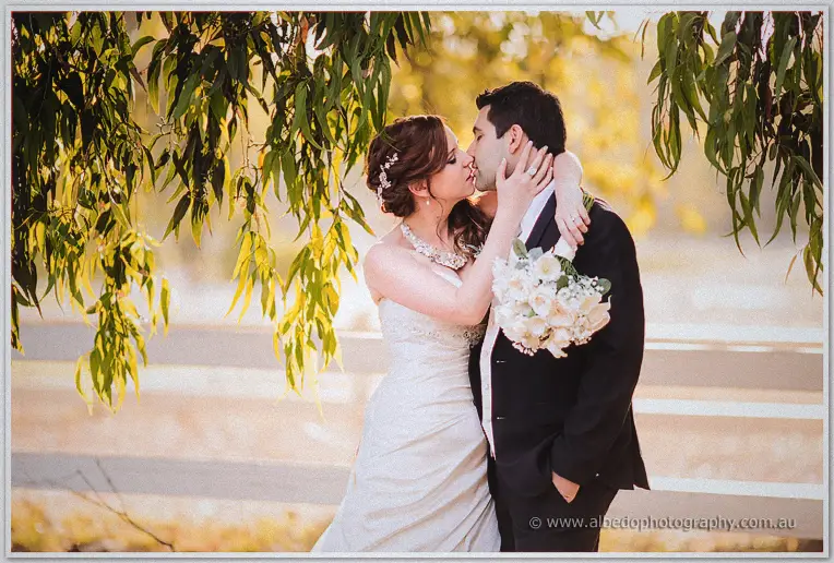 Amanda cradles Craig's face for a kiss under sunlit gum branches by the paddock