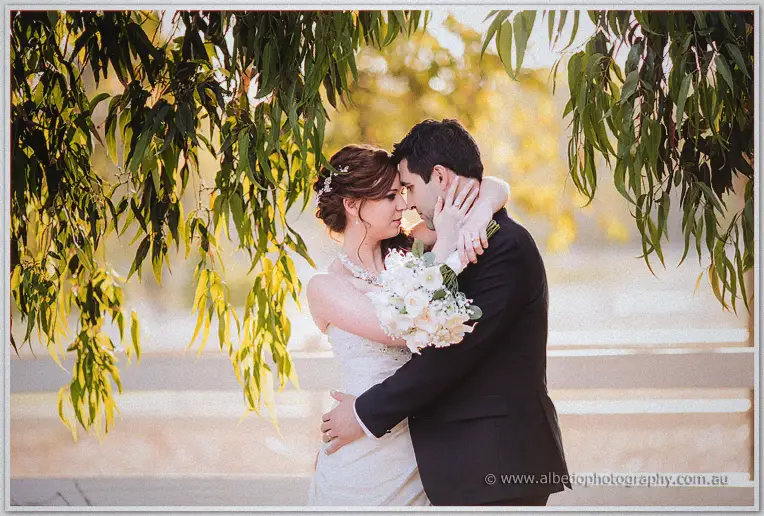 Bride and groom press foreheads together, her bouquet tucked between them under leaves