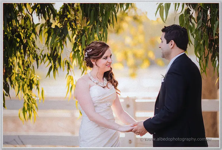 Amanda smiles across at Craig as they hold hands beneath golden foliage and white rails