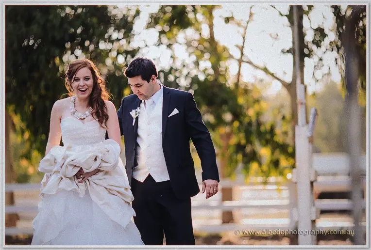 The couple laugh mid-stride along the paddock fence in soft evening light