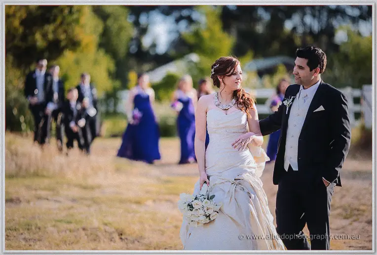 Amanda and Craig stroll toward the camera, wedding party trailing in sunlit blur