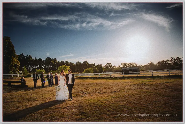 Amanda and Craig cross a wide golden paddock, party behind them under a vast dusk sky