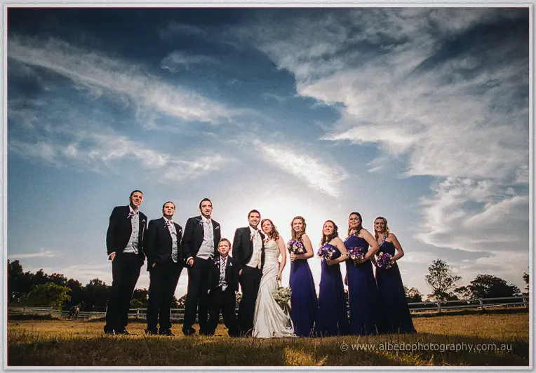 Bridal party silhouetted against dramatic clouds, sun flaring behind the couple