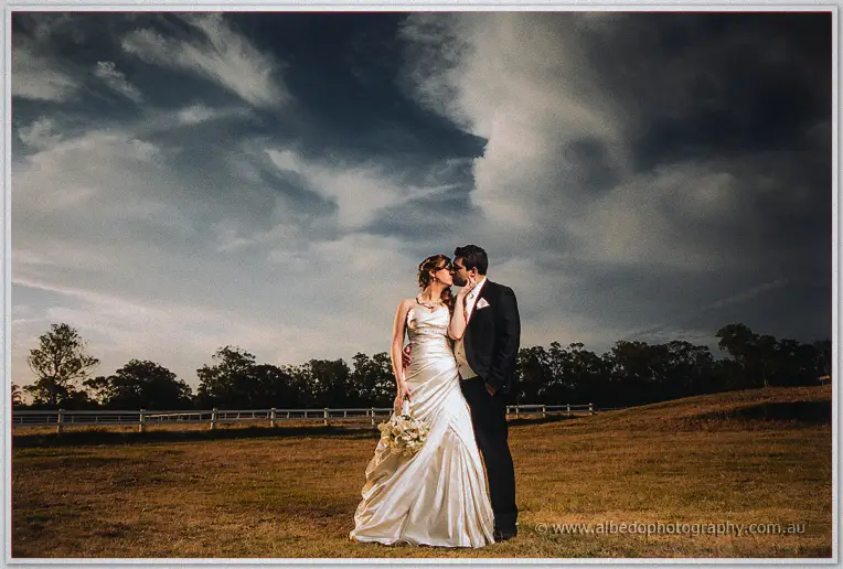 Amanda and Craig kiss beneath a dramatic cloudy sky in a Swan Valley paddock ringed by white fencing