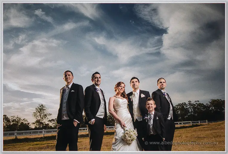Craig's groomsmen and a cheeky page boy flank Amanda in her gown beneath sweeping evening clouds at Brookleigh