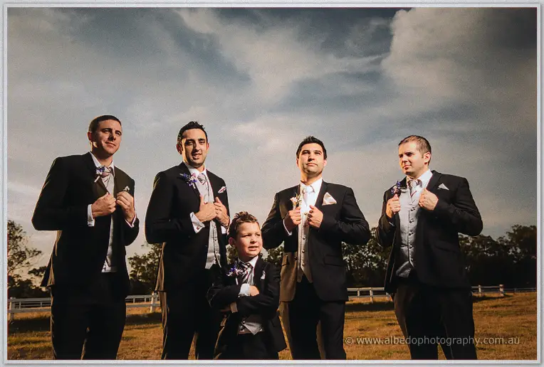 Groomsmen straighten their jackets around a confident page boy with crossed arms under a moody country sky