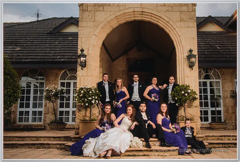 The full bridal party pose on the sandstone steps of Brookleigh's arched entry, purple gowns glowing at dusk