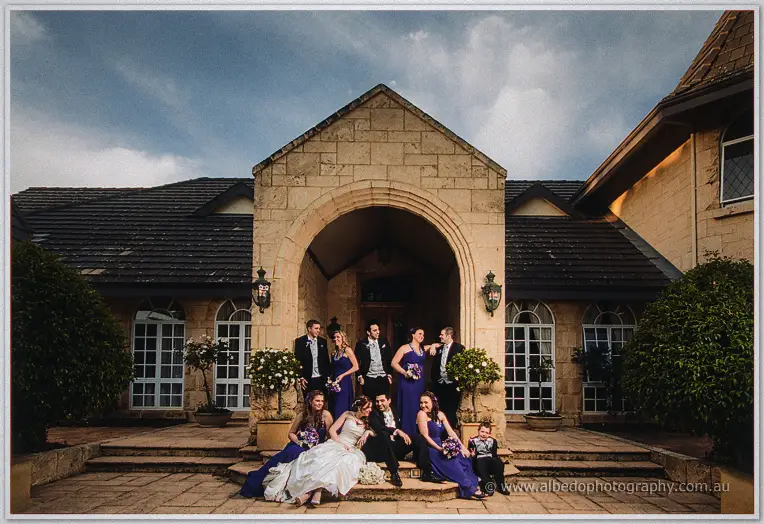 Bridesmaids in purple and groomsmen in black gather on the steps beneath the stone arch entrance