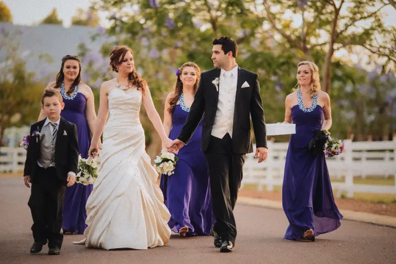 Craig leads Amanda down a jacaranda-lined drive with their purple-clad bridal party in golden light
