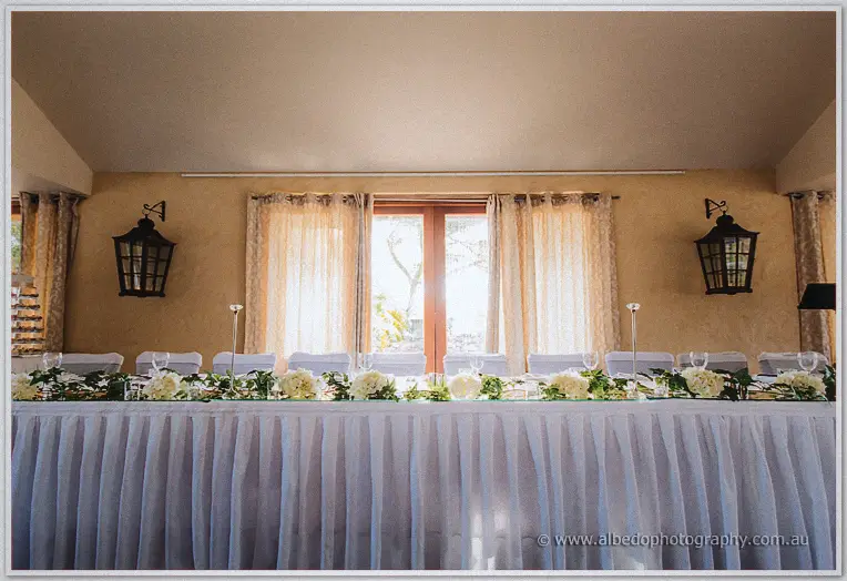 The bridal table in white with rose garlands glows warmly between twin lanterns and tall curtained windows