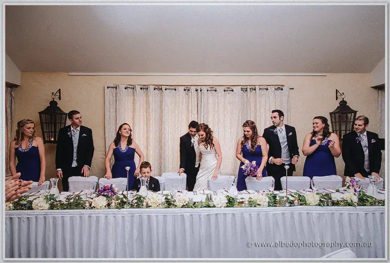 The wedding party assembles behind the bridal table under lantern sconces and sheer white drapes