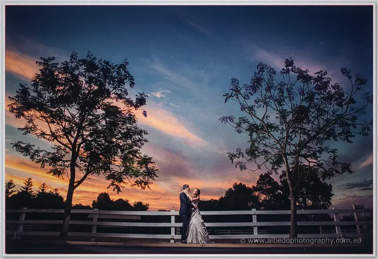 A wide sunset vista frames Amanda and Craig kissing on the gravel between silhouetted jacaranda trees