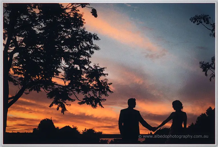 Silhouettes of the couple holding hands burn orange against the fiery sunset sky at Brookleigh Estate