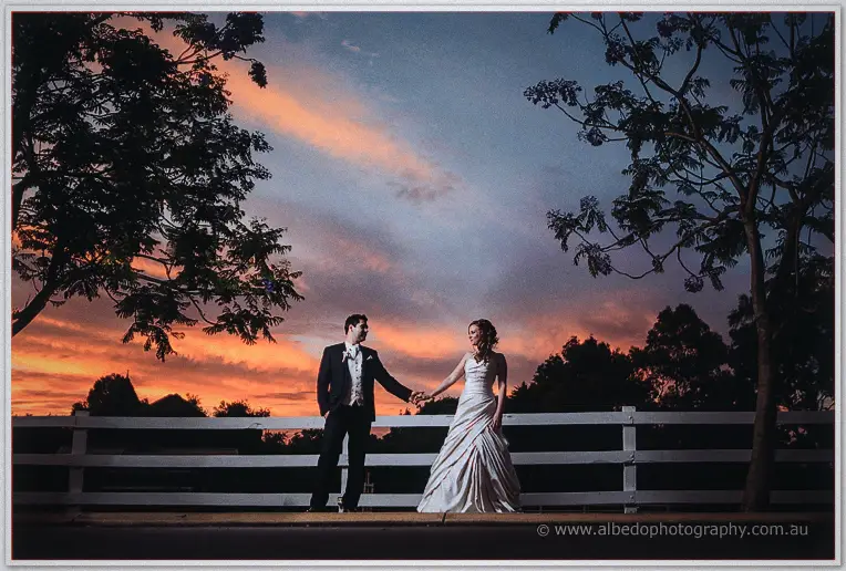 Hand in hand before the white fence, Amanda and Craig pause as peach light washes over the estate grounds