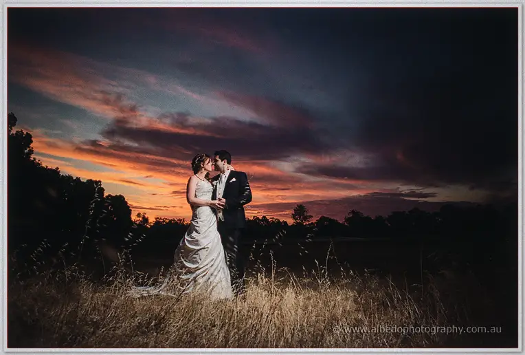 Amanda and Craig embrace in a golden field beneath a fiery sunset sky streaked with orange and violet