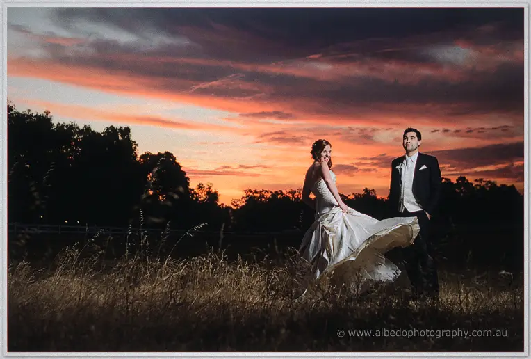 Amanda lifts her gown and laughs towards Craig in the tussocky paddock under deep red evening cloud