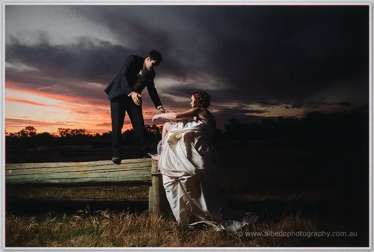 Craig reaches down from the timber fence rail to help Amanda up as storm clouds swallow the sunset