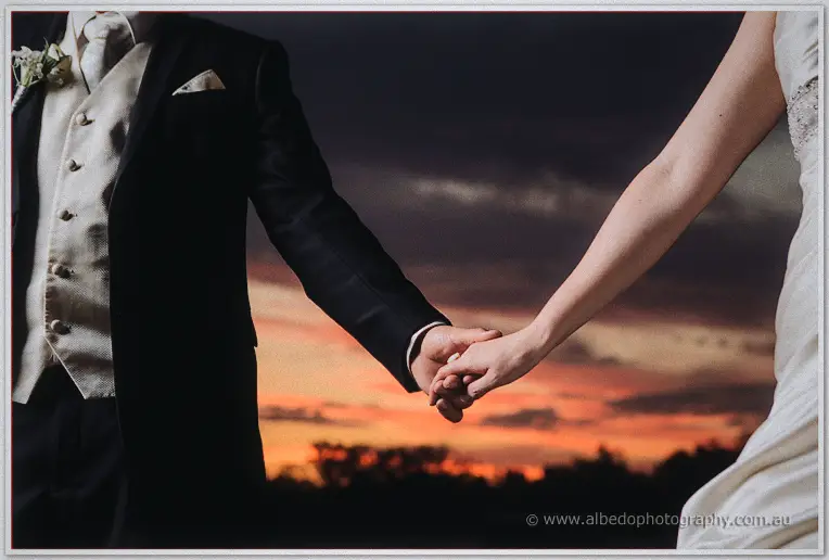 Craig in a black suit and cream waistcoat holds Amanda's hand against a fiery sunset sky