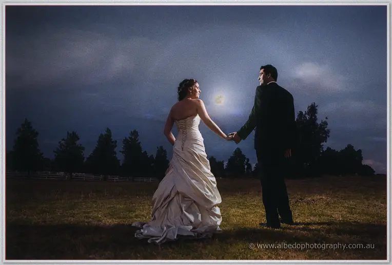 Amanda and Craig walk hand in hand across the dusk-lit paddock with the moon glowing faintly behind them