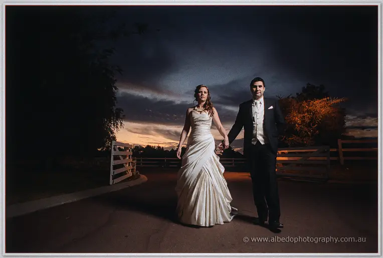 Amanda and Craig walk the drive hand in hand under a night sky, lantern-lit trees glowing warm behind them