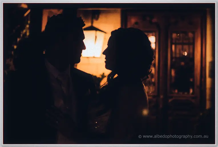 The couple's profiles meet in silhouette against glowing lamplight at the warmly lit homestead entrance