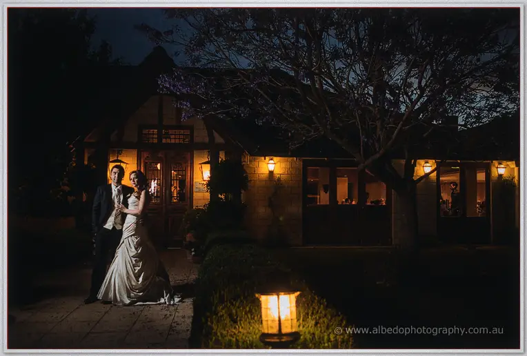 Amanda and Craig stand close outside the floodlit stone homestead at night, a garden lantern glowing nearby