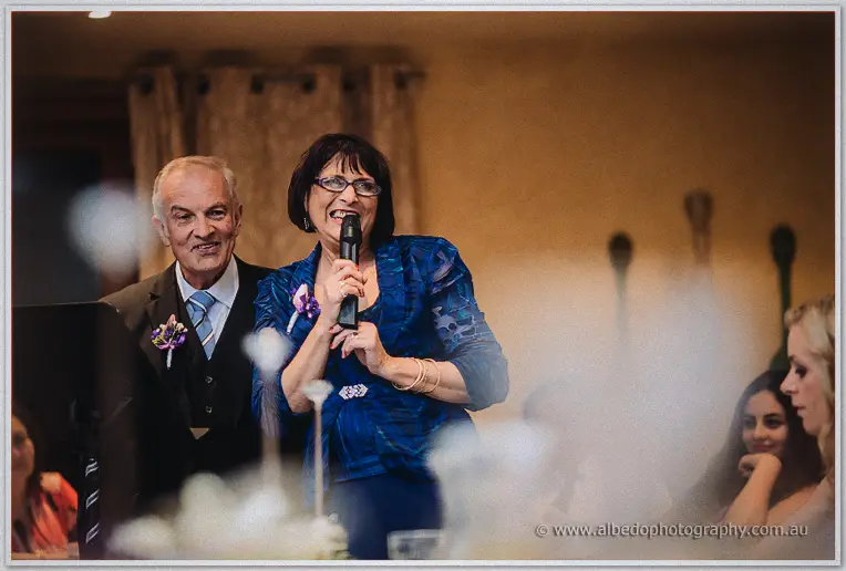 A laughing guest in a cobalt jacket delivers her toast beside a beaming gent at the reception