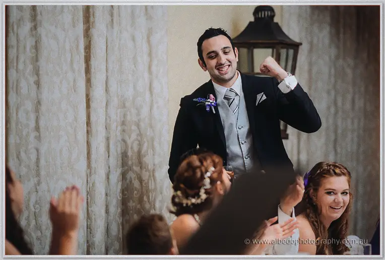 A groomsman pumps his fist mid-speech while bridesmaids laugh and guests applaud below the wall lantern