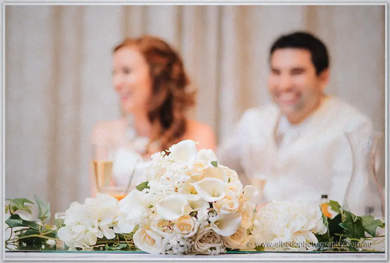 A blush bouquet of calla lilies and roses sits sharp in front while Amanda and Craig laugh softly behind