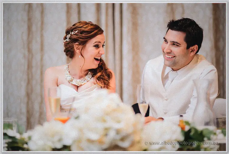 Amanda gasps in delight beside a grinning Craig as white blooms cascade across the bridal table