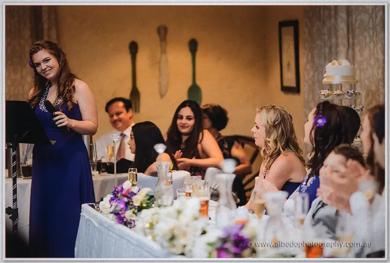 A bridesmaid in royal purple addresses the room from the lectern as smiling guests listen near the cake