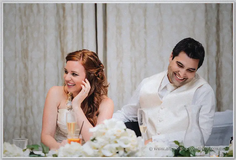 Amanda laughs with a hand to her ear while Craig bows his head chuckling behind the flower garland