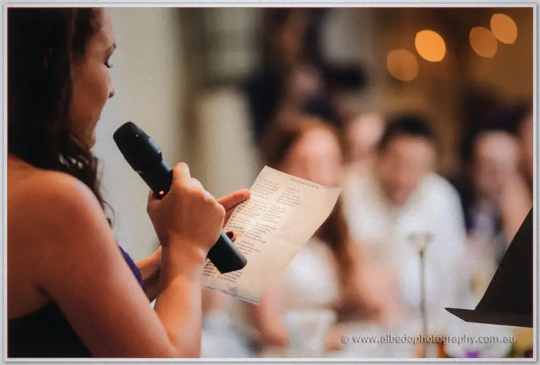 A bridesmaid grips the microphone and her speech notes while blurred guests watch through the bokeh