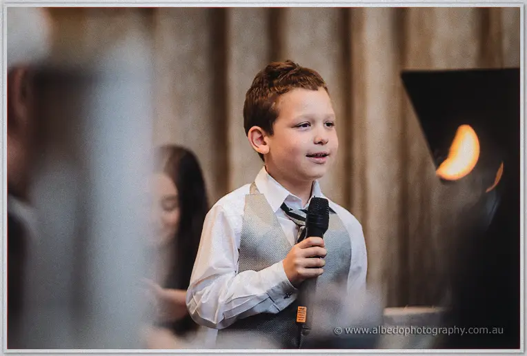 The young page boy in a grey vest and tie speaks earnestly into a microphone taller than his chin