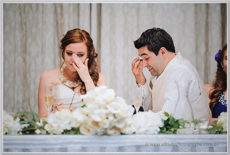 Amanda dabs her tears with a tissue as Craig wipes his eye behind the white roses at the bridal table