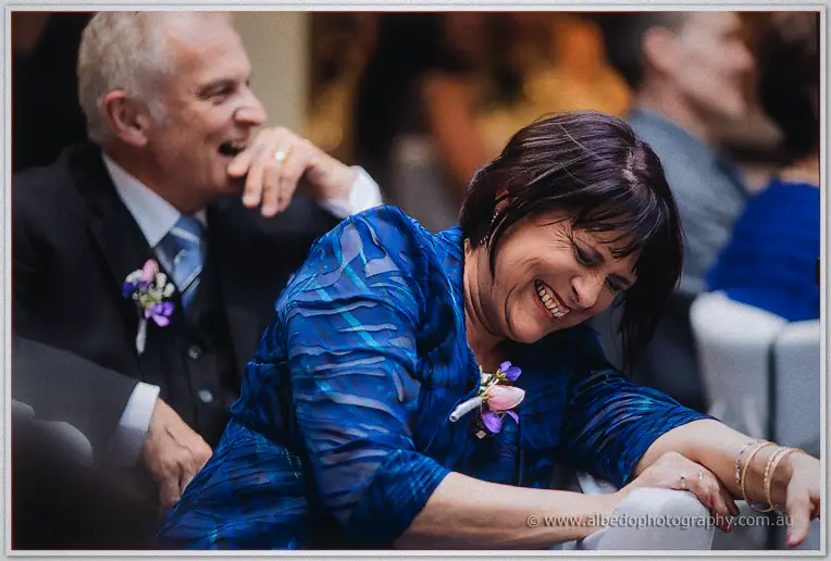 A guest in shimmering blue doubles over laughing beside a chuckling gentleman in a suit at the reception